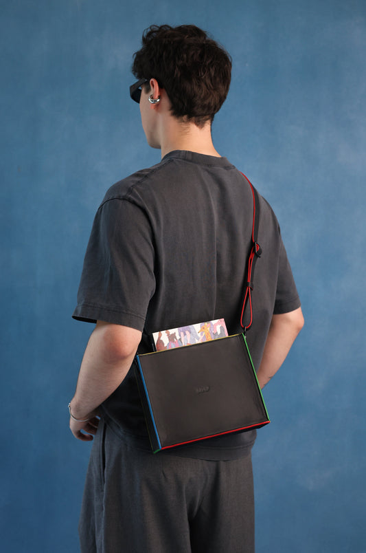 A person shown from the back wearing a dark gray t-shirt and shorts, carrying a black crossbody bag with a red strap and colorful edges over their shoulder, set against a textured blue studio backdrop.