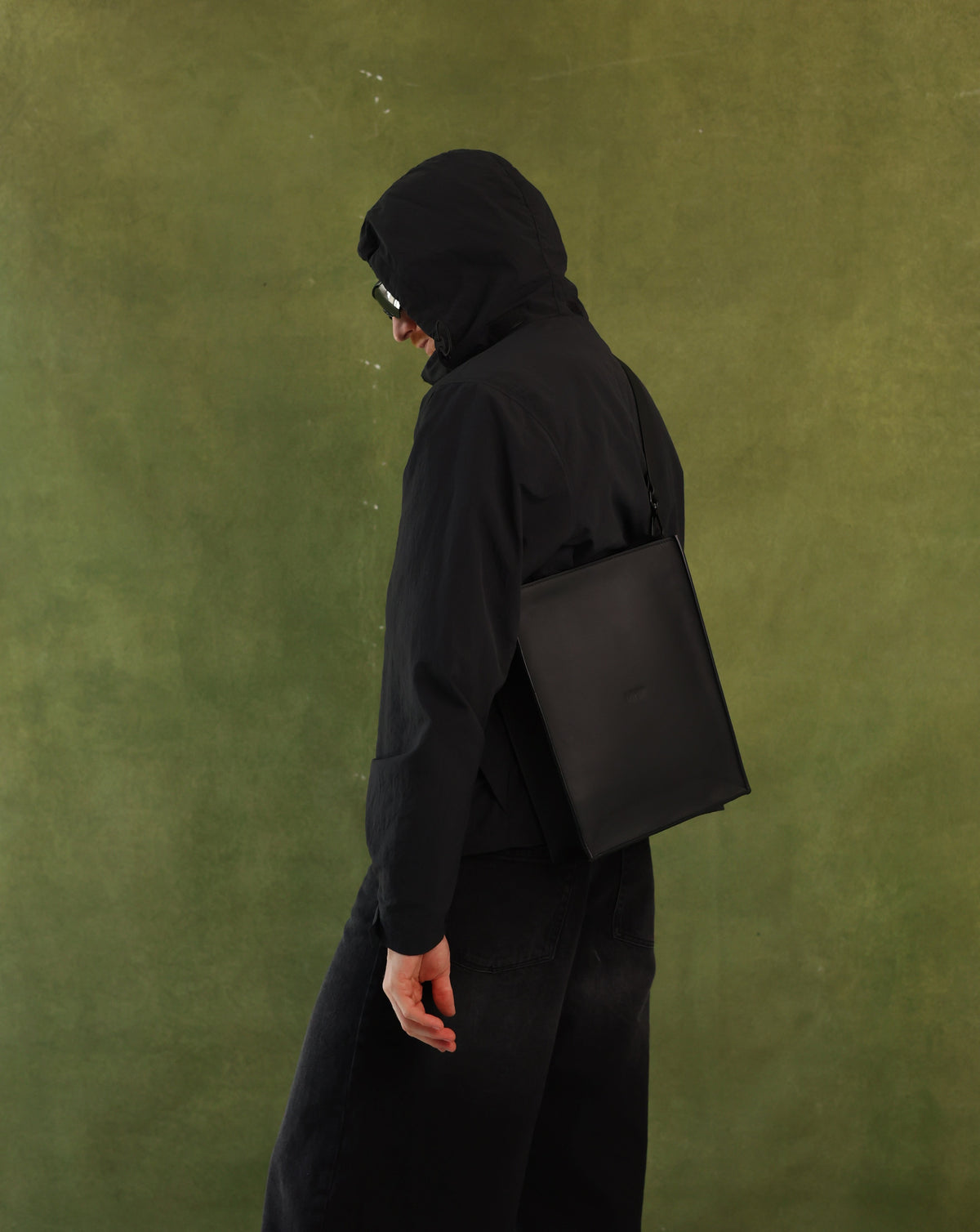 A person in a black hooded jacket and black pants, shown from the back and side, carrying a black leather shoulder bag against a textured olive green background.