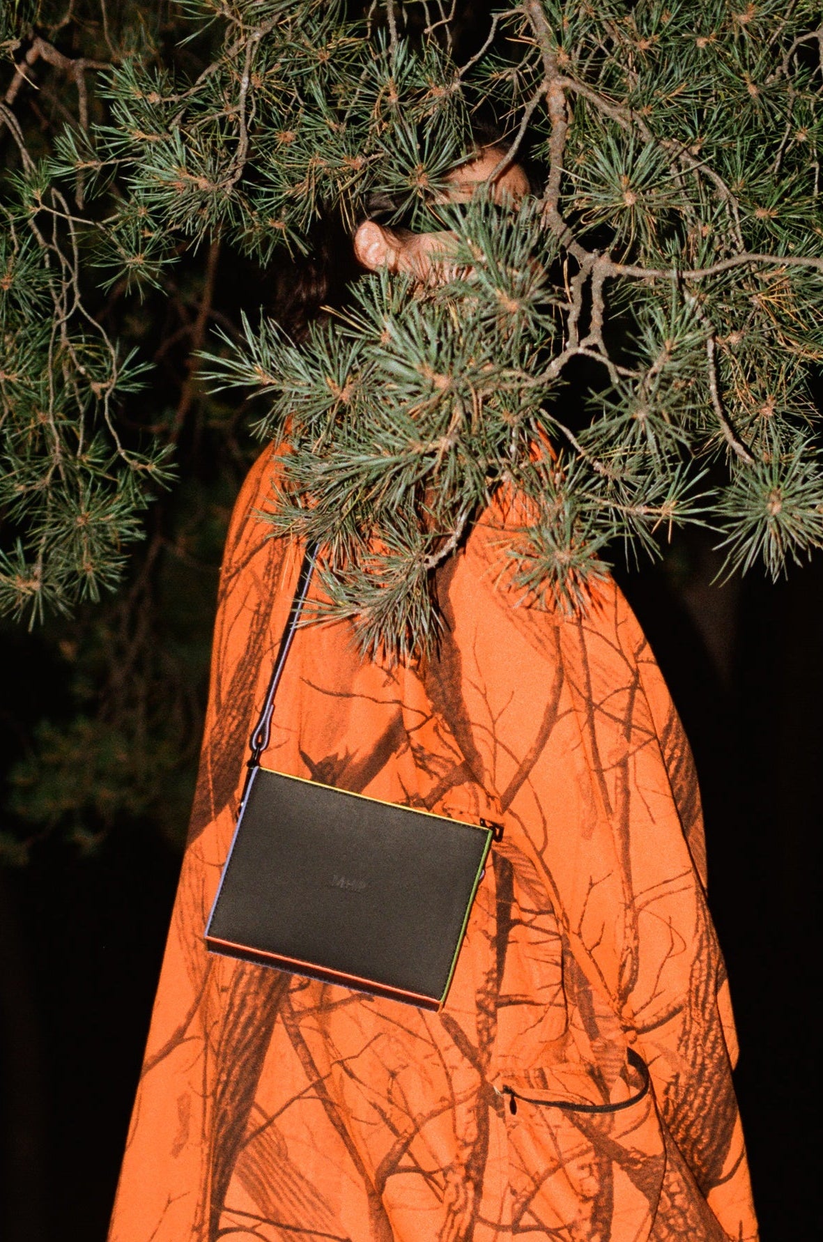 A person standing outdoors at night under pine branches, wearing an orange dress with a dark abstract branch-like print, tan boots, and carrying a black shoulder bag with colored edges. Which are purple, orange, green and yellow.