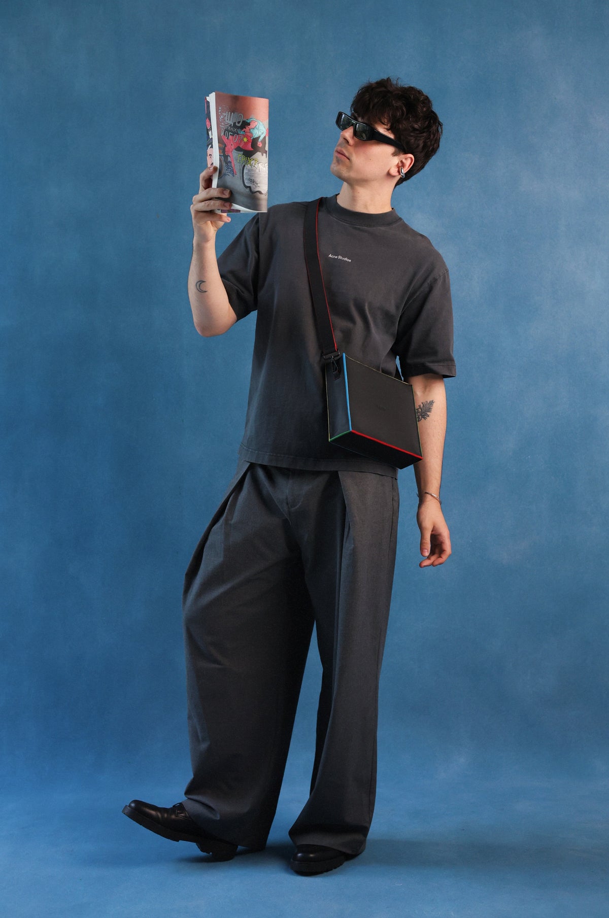 A man wearing a dark gray t-shirt, matching wide-leg trousers, and sunglasses holds a magazine while wearing a black crossbody bag with red trim and a blue side accent, set against a textured blue studio backdrop.