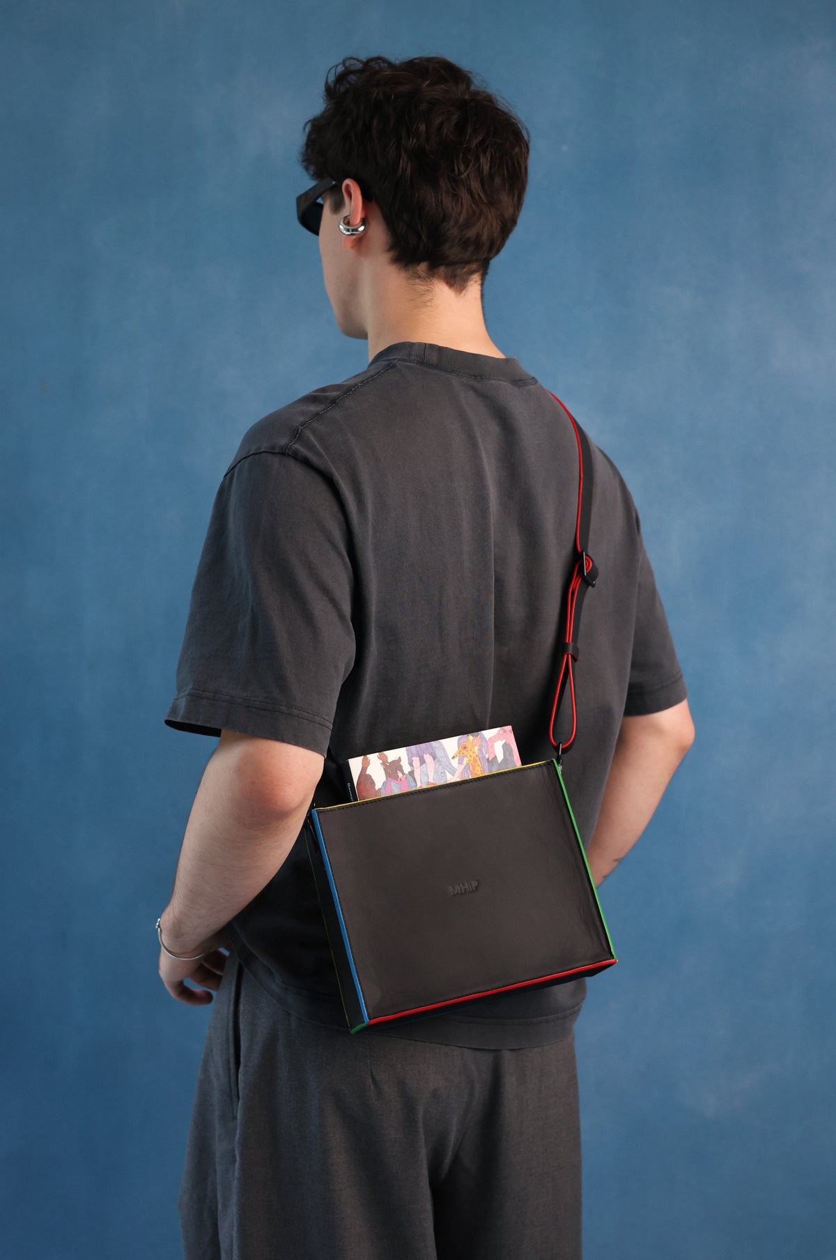 A person shown from the back wearing a dark gray t-shirt and shorts, carrying a black crossbody bag with a red strap and colorful edges over their shoulder, set against a textured blue studio backdrop.