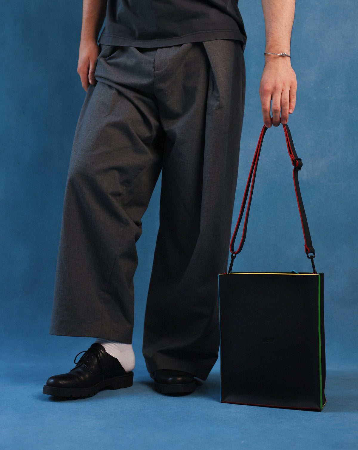 A person wearing loose dark gray wide-leg trousers, a dark gray t-shirt, white socks, and black leather shoes holds a black tote bag with red and green trim and a red shoulder strap against a blue studio backdrop.