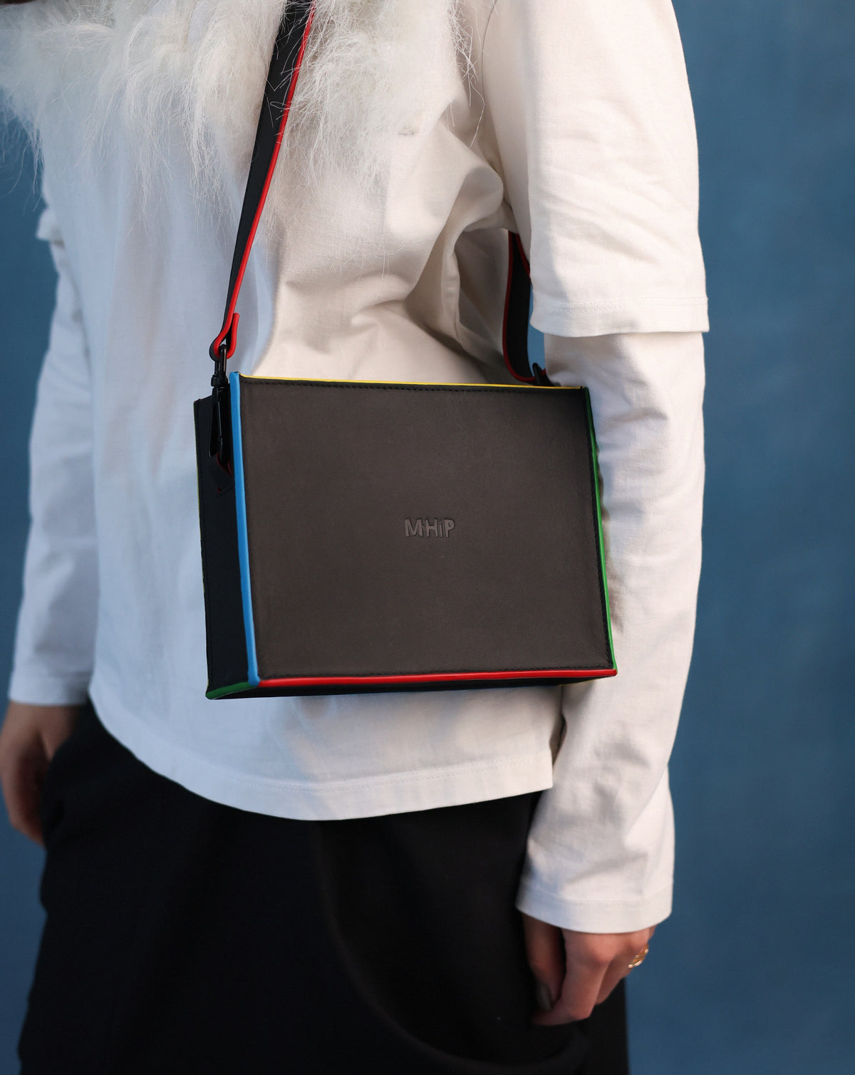 A person wearing a white long-sleeve top and black skirt carries a structured black crossbody bag with a thin multicolor striped edge and a black strap with red and blue accents. The bag has the letters 'MHP' printed on the front.