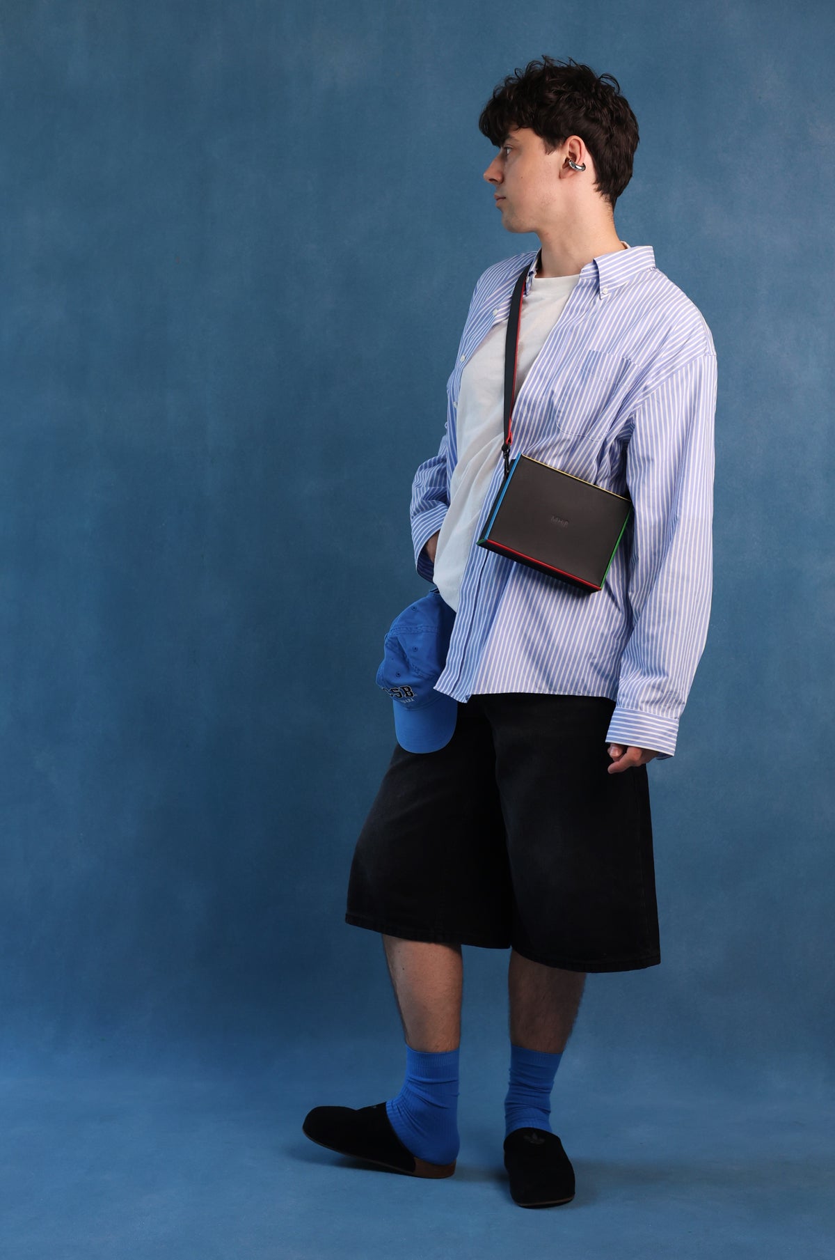 A person stands in profile against a blue studio backdrop, wearing a light blue and white vertically striped button-up shirt, black shorts, blue socks, and black slip-on shoes. A dark rectangular bag with a red accent is slung over the shoulder, and a blue cap hangs from the pocket.
